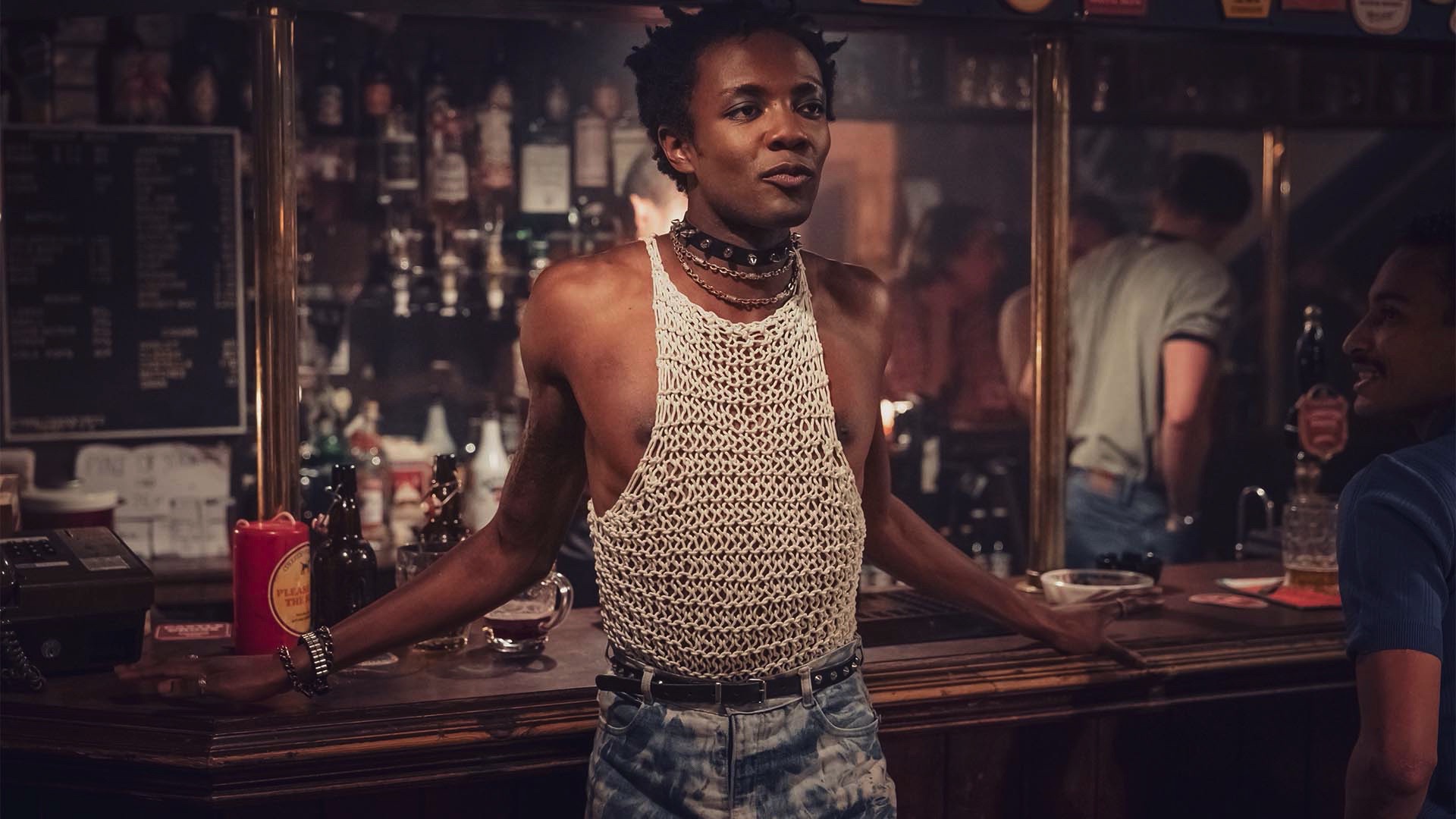 Omari Douglas stands in a bar in a string vest as Roscoe Babatunde
