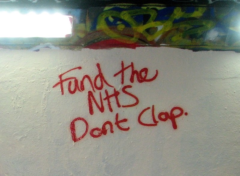 Red writing on a white wall which reads "Fund the NHS. Don't clap".