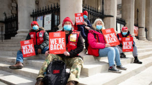 Five vendors sitting on the steps of St Martin In the Fields wearing PPE masks and holding magazine's saying "We're back"