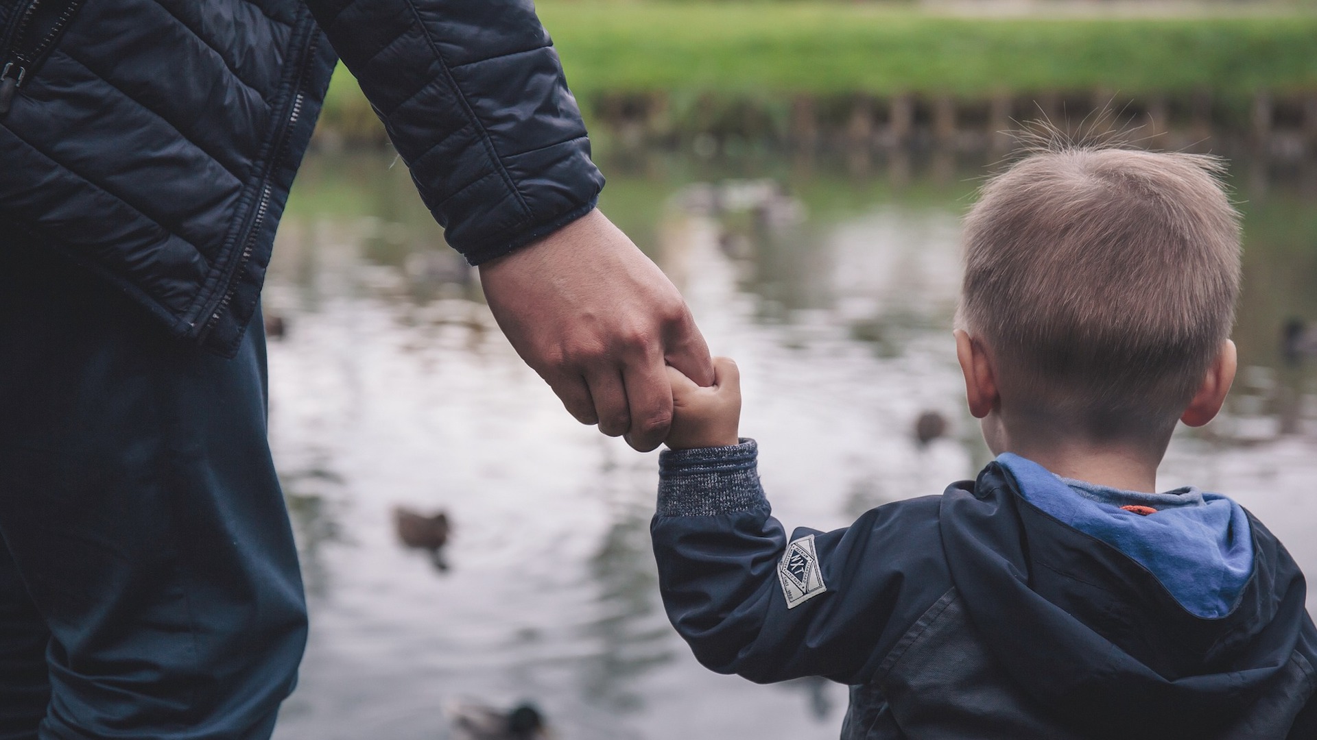Adult holding a child's hand in front of a pond. child poverty