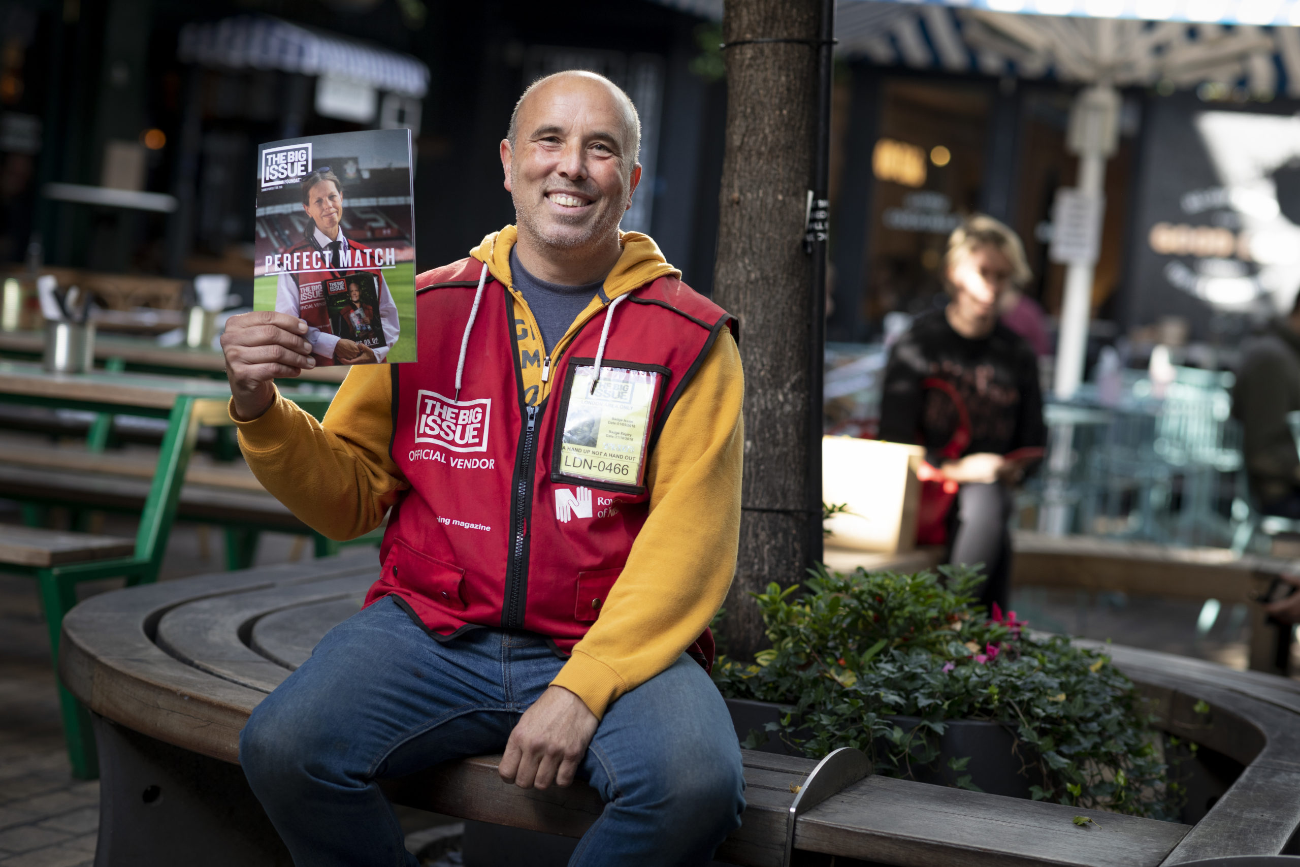 Big Issue vendor holding up issue of magazine, asking for regular donations to The Big Issue Foundation.