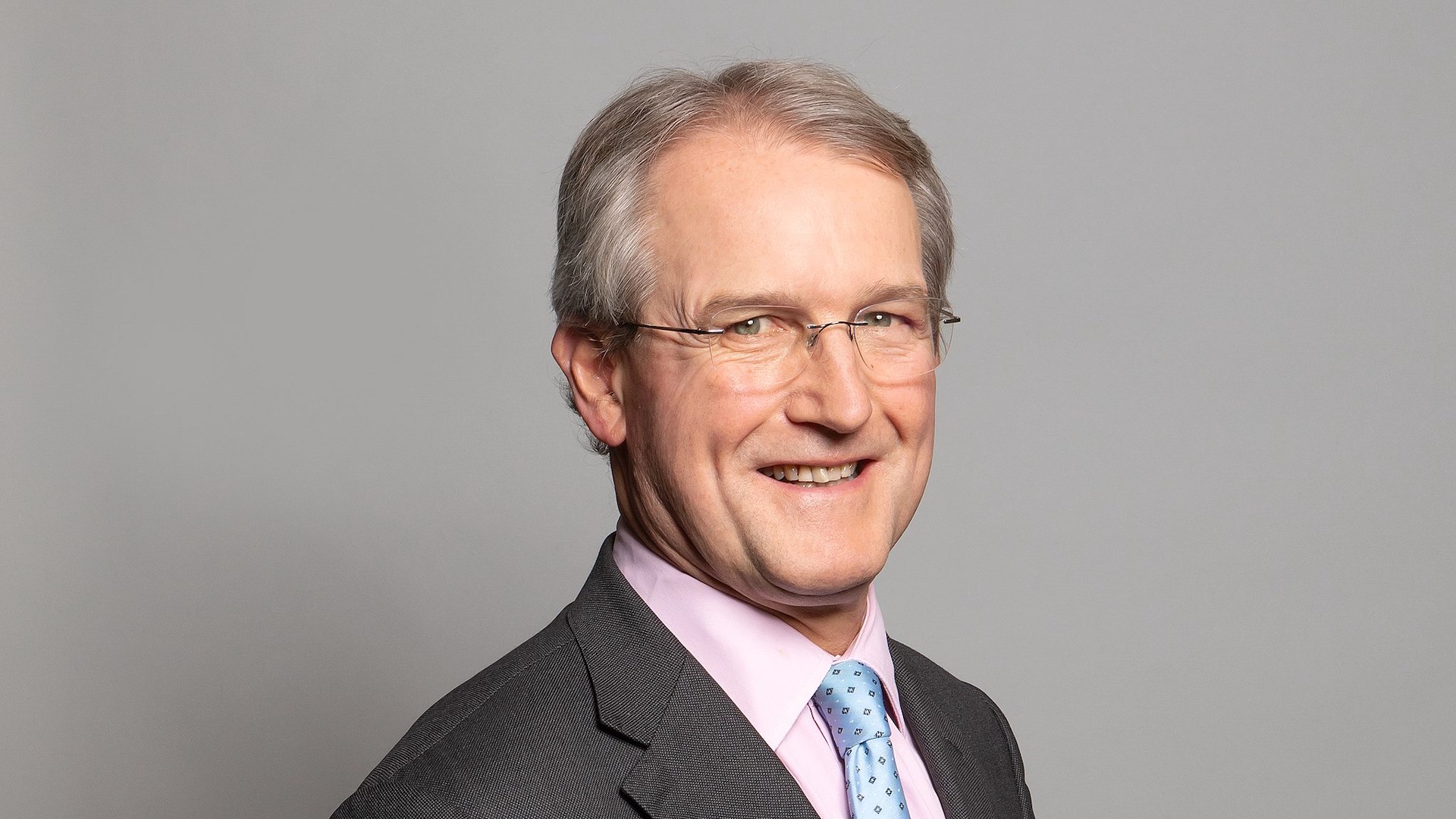 Owen Paterson's official portrait, wearing glasses a pink tie and blue shirt