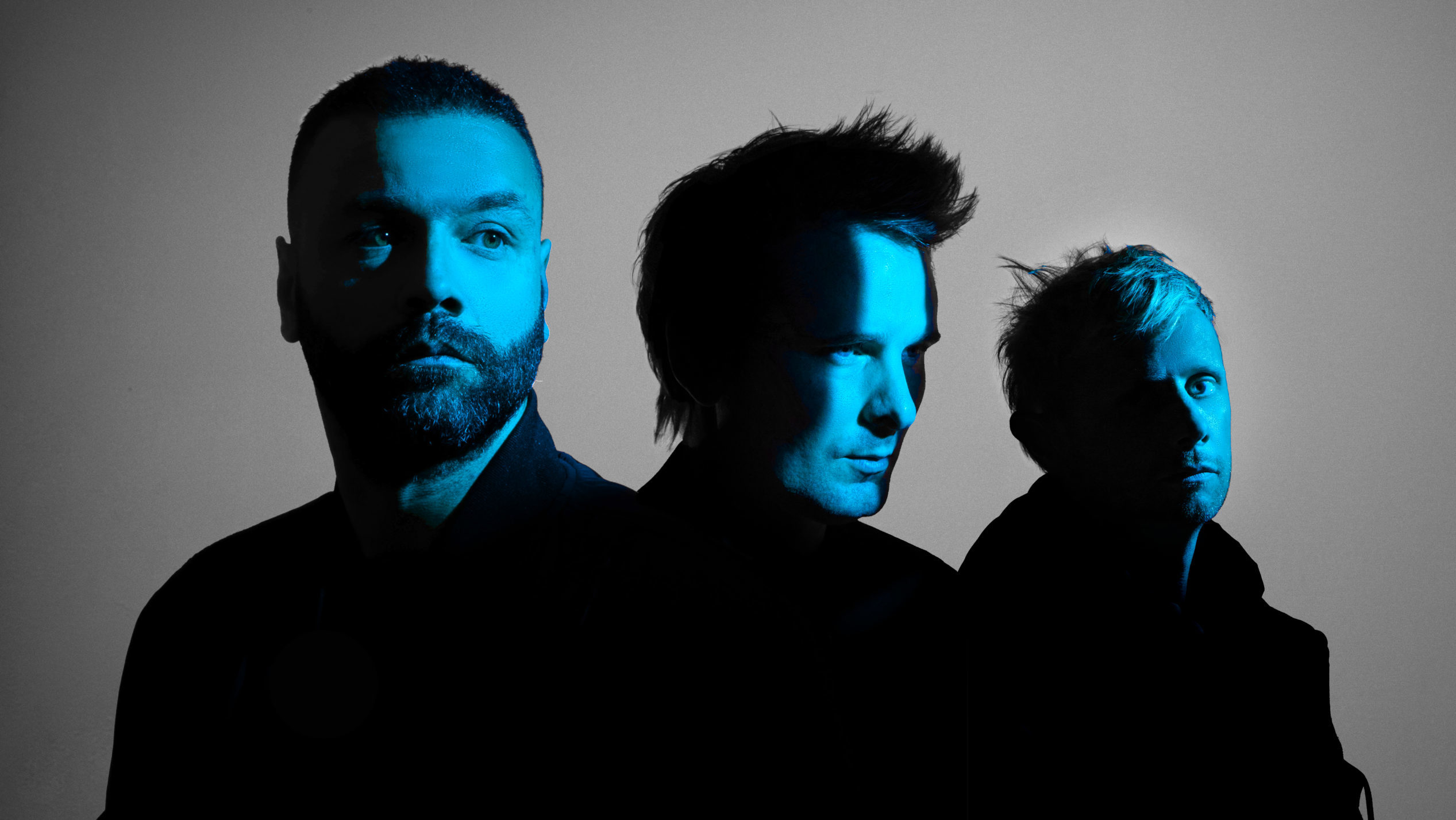 Muse's Matt Bellamy, Chris Wolstenholme, Dominic Howard stand in darkness except for blue light reflected from their faces