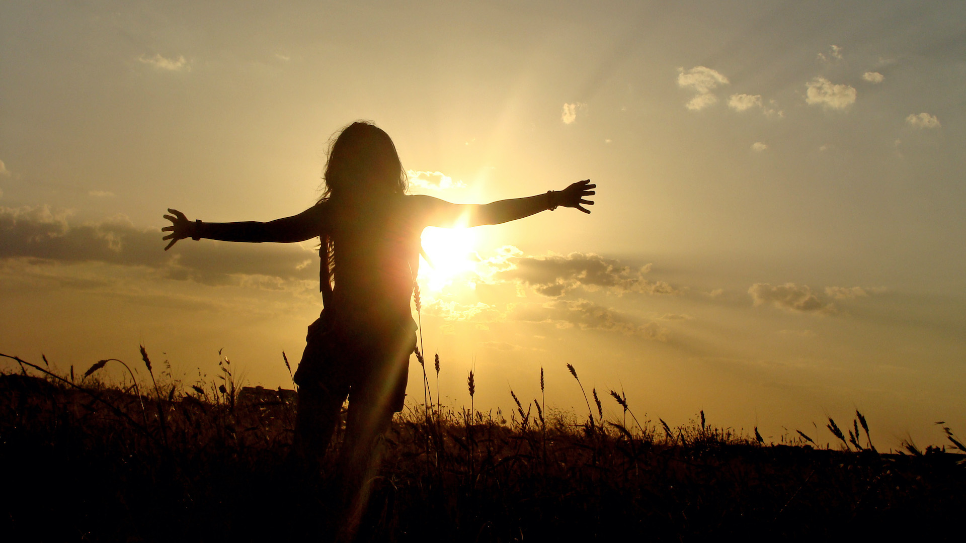 A woman stands in a field of grass, silhouetted by the sun