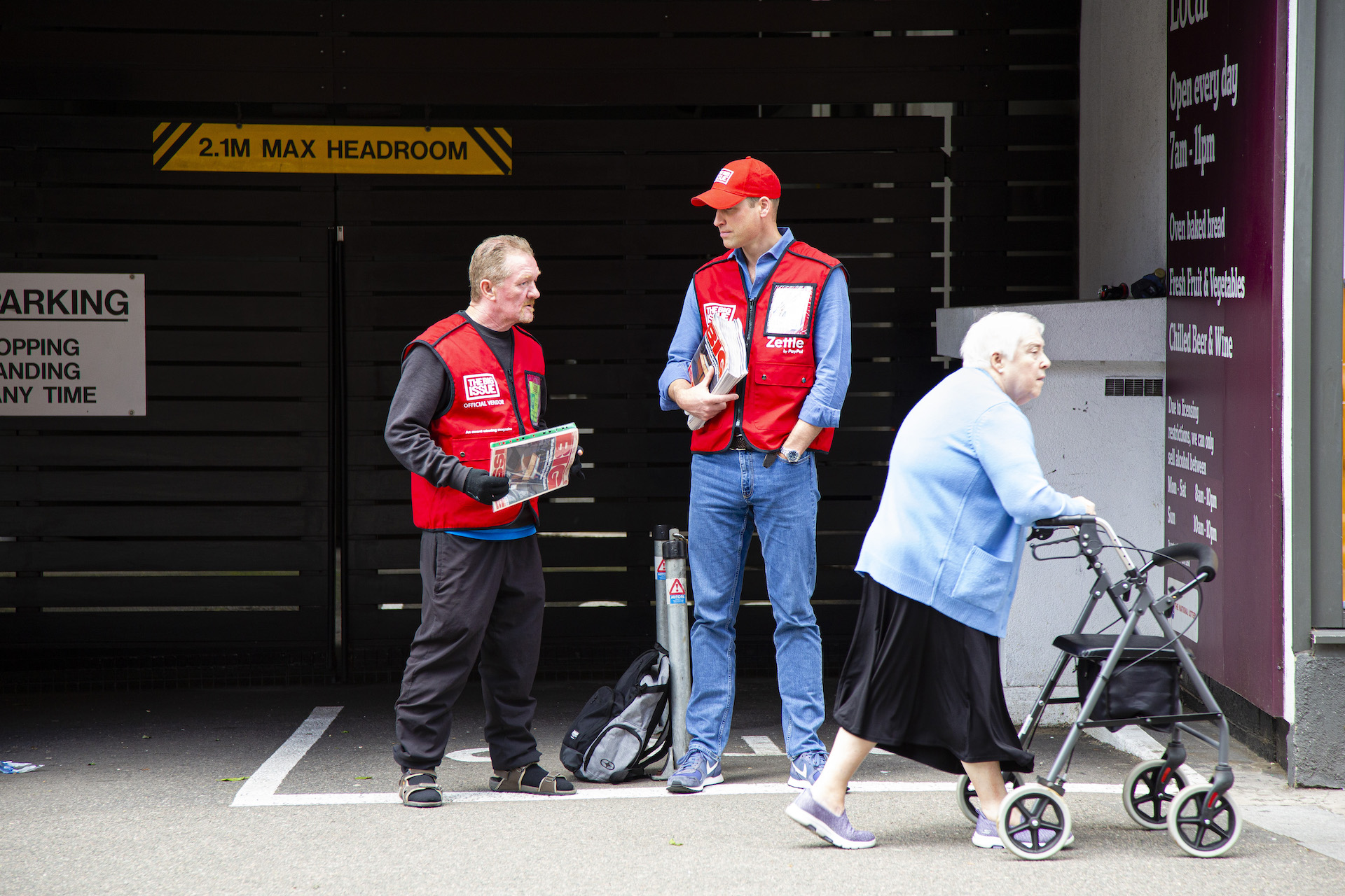 Prince WIlliam and Big Issue seller Dave Martin in red Big Issue hats and tabards stand in conversation as an elderly woman walks past