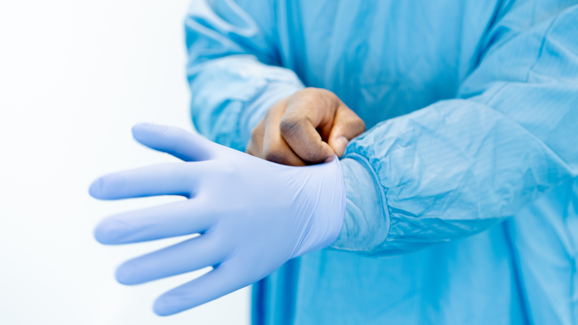 A man pulls a blue glove on to his left hand while wearing a medical gown