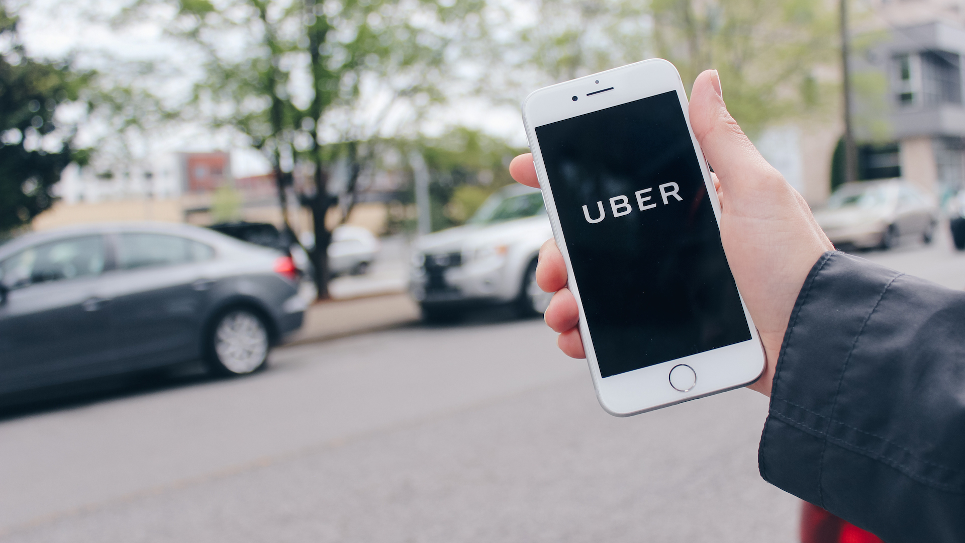 A hand comes in from the side of the screen holding a mobile phone with the Uber app open. Cars are out of focus in the background