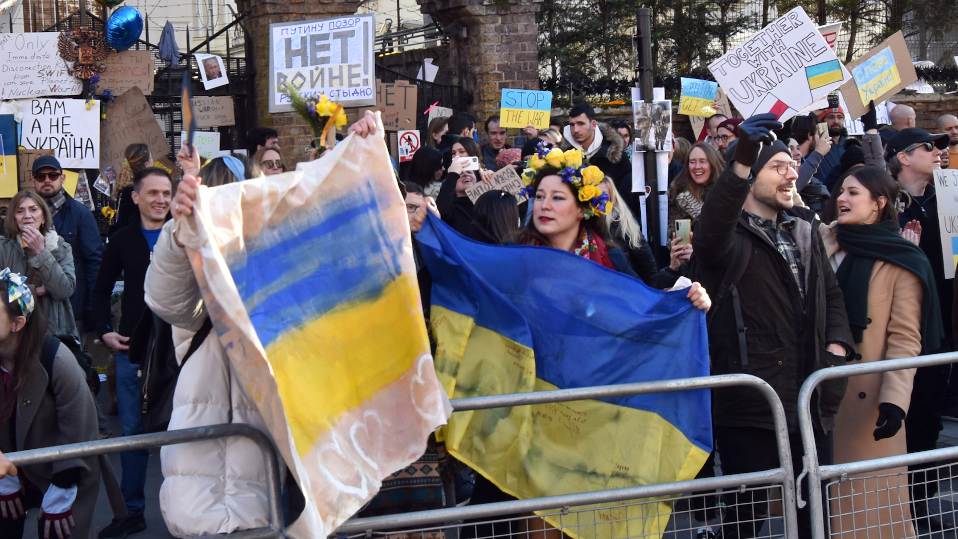Crowds hold Ukrainian flags and signs in front of barriers at the Russian Embassy in London