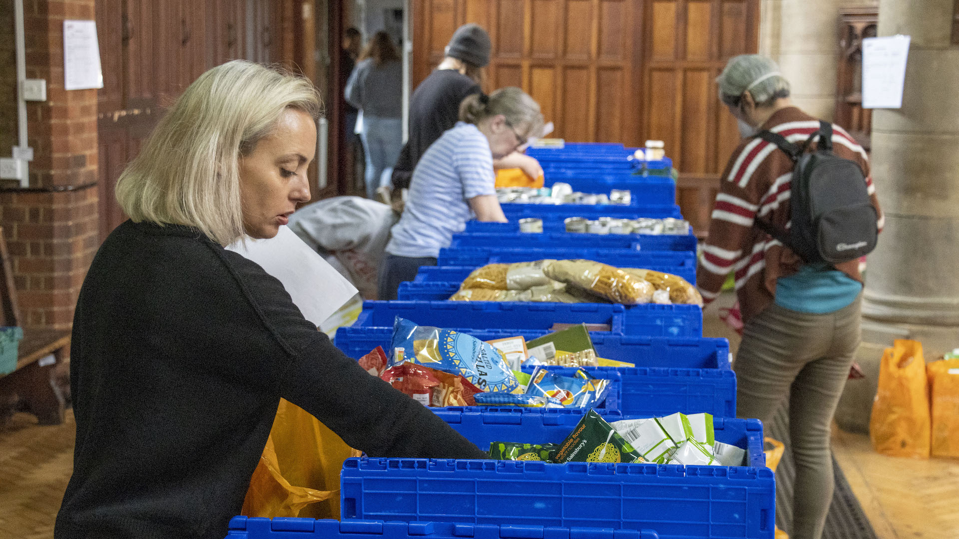 A woman at a food bank reaches into a blue plastic container filled with food