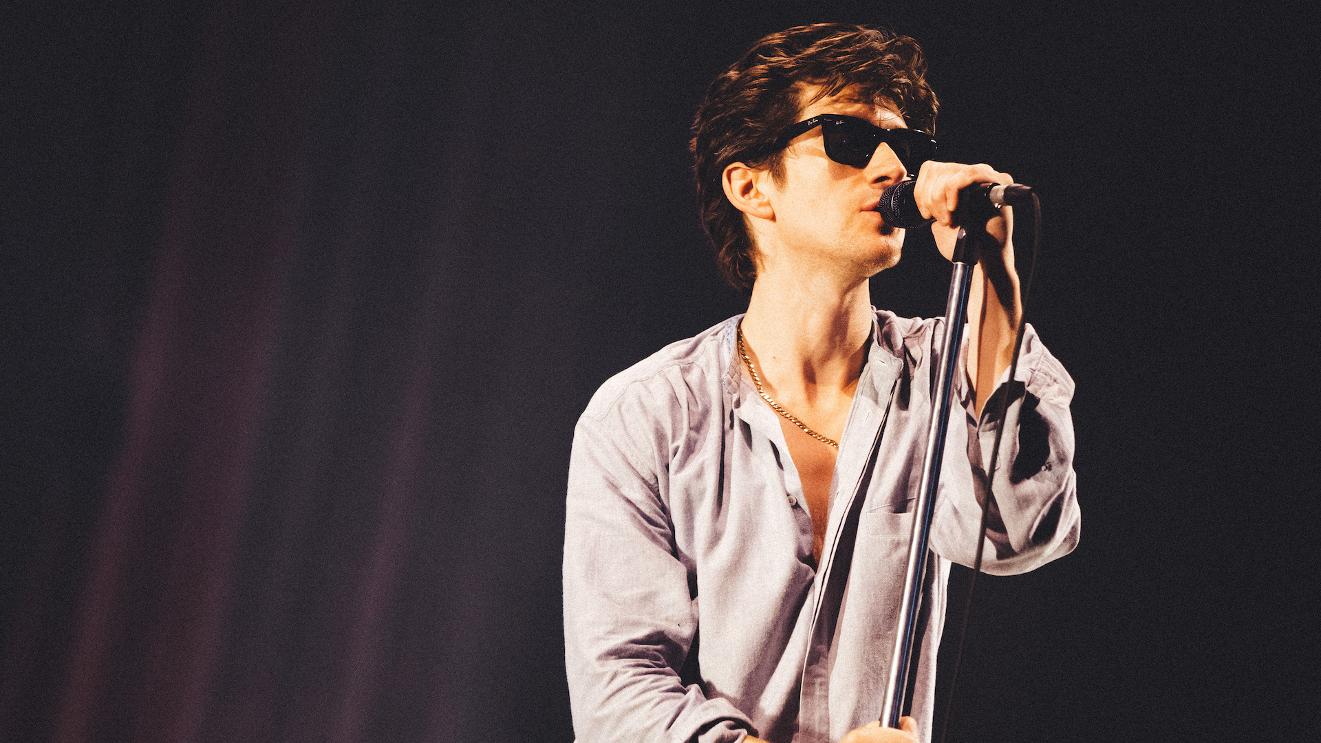 arctic monkeys singer alex turner leans into a microphone while singing and wearing an open pale blue shirt and sunglasses