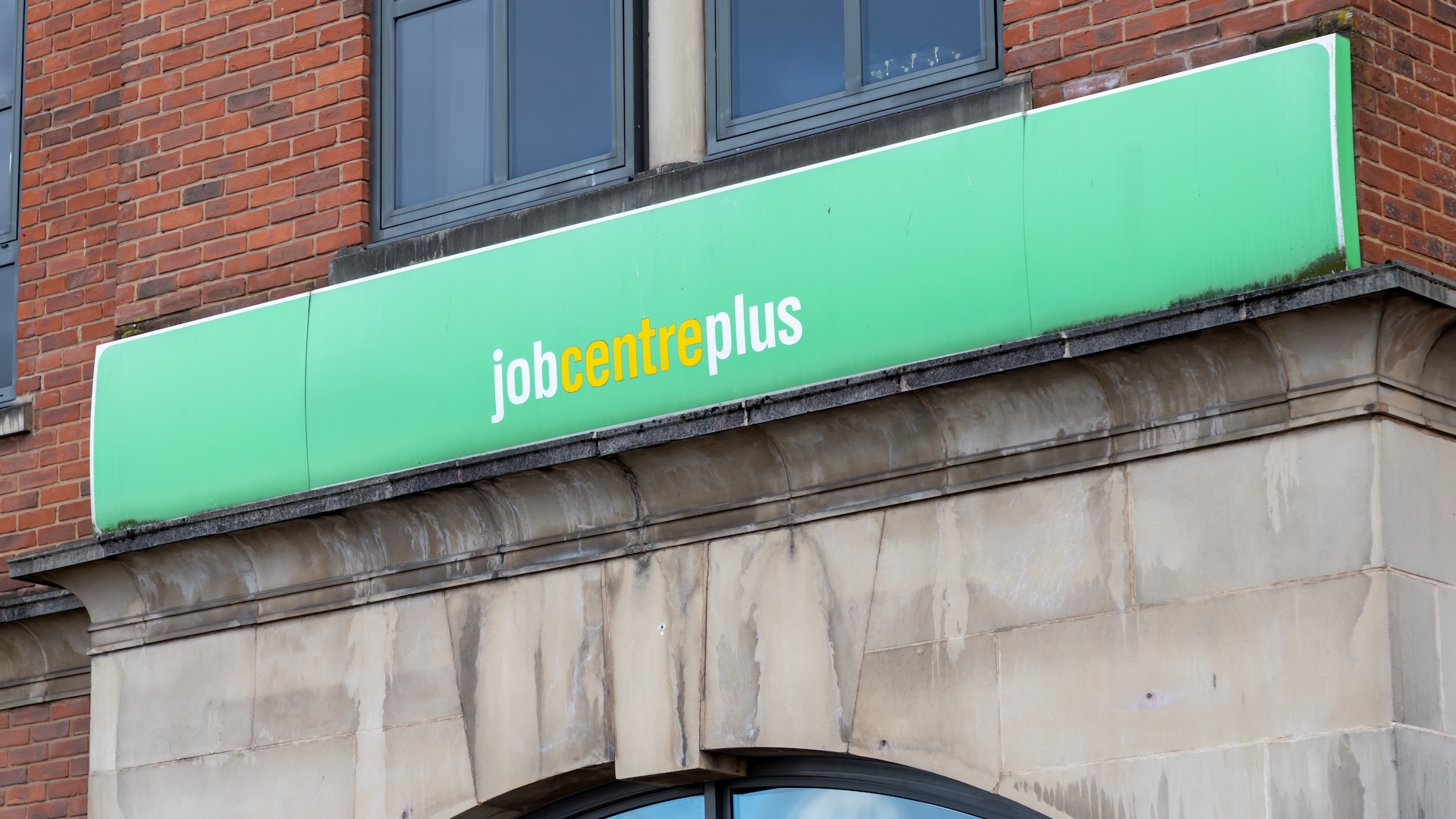 The green job centre plus sign hangs above a wet and dirty concrete architrave