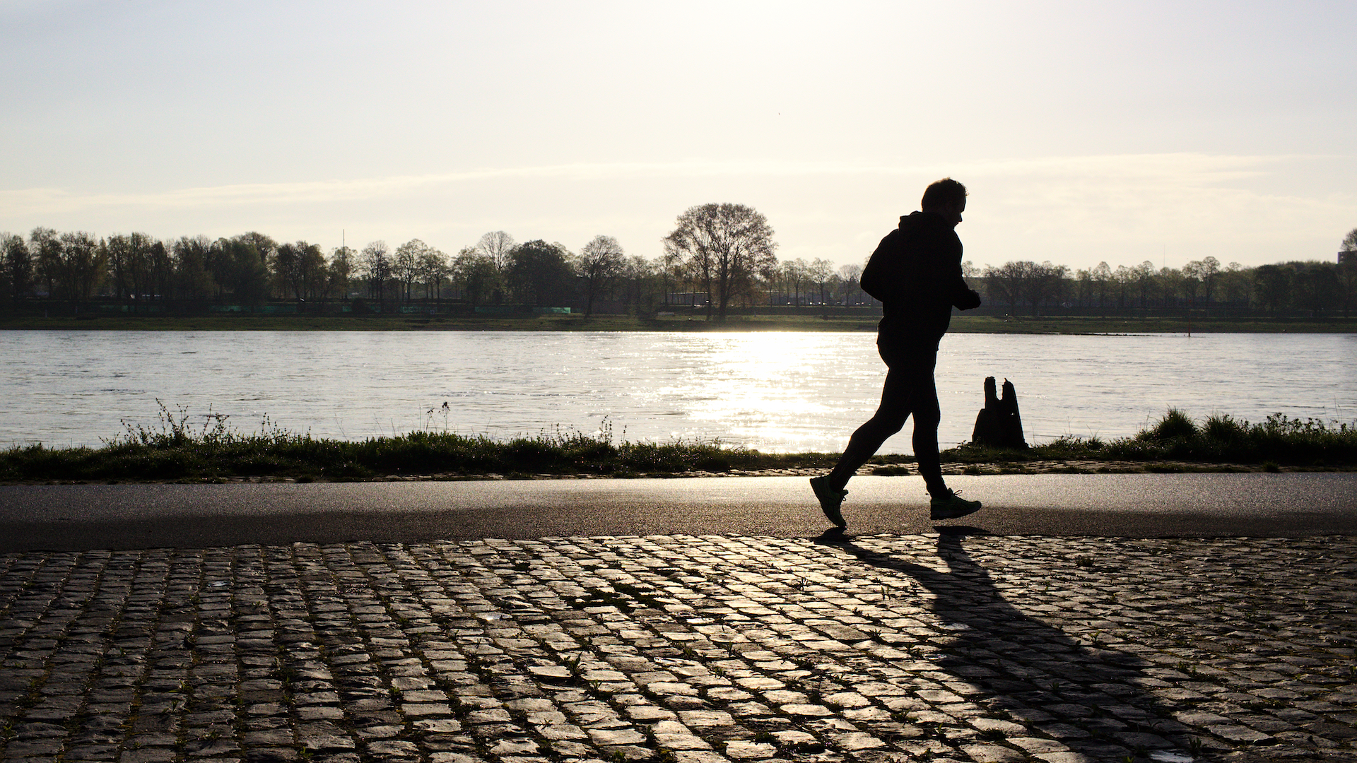 A silhouetted man runs on cobblestones alongside a river in winter
