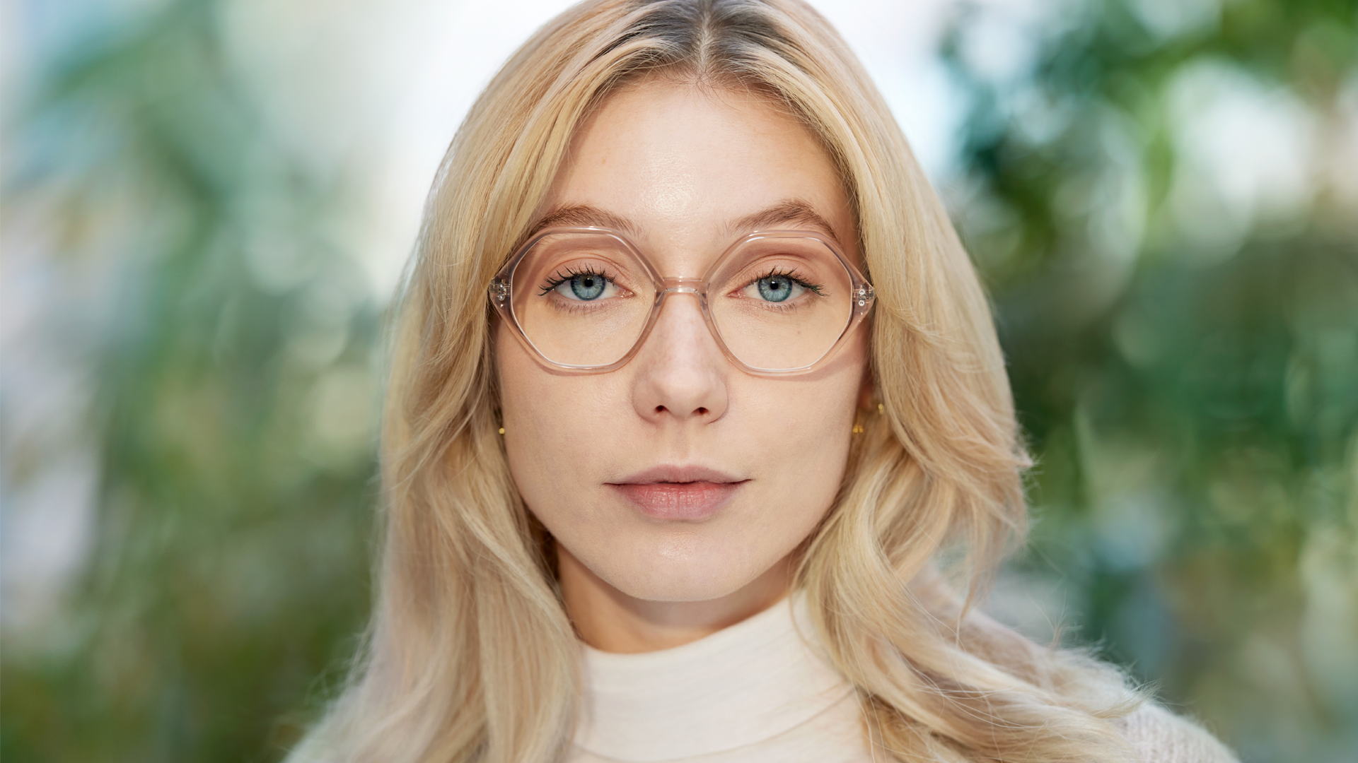 A blonde woman wearing fashionable spectacles with transparent frames, against a backdrop to out-of-focus trees