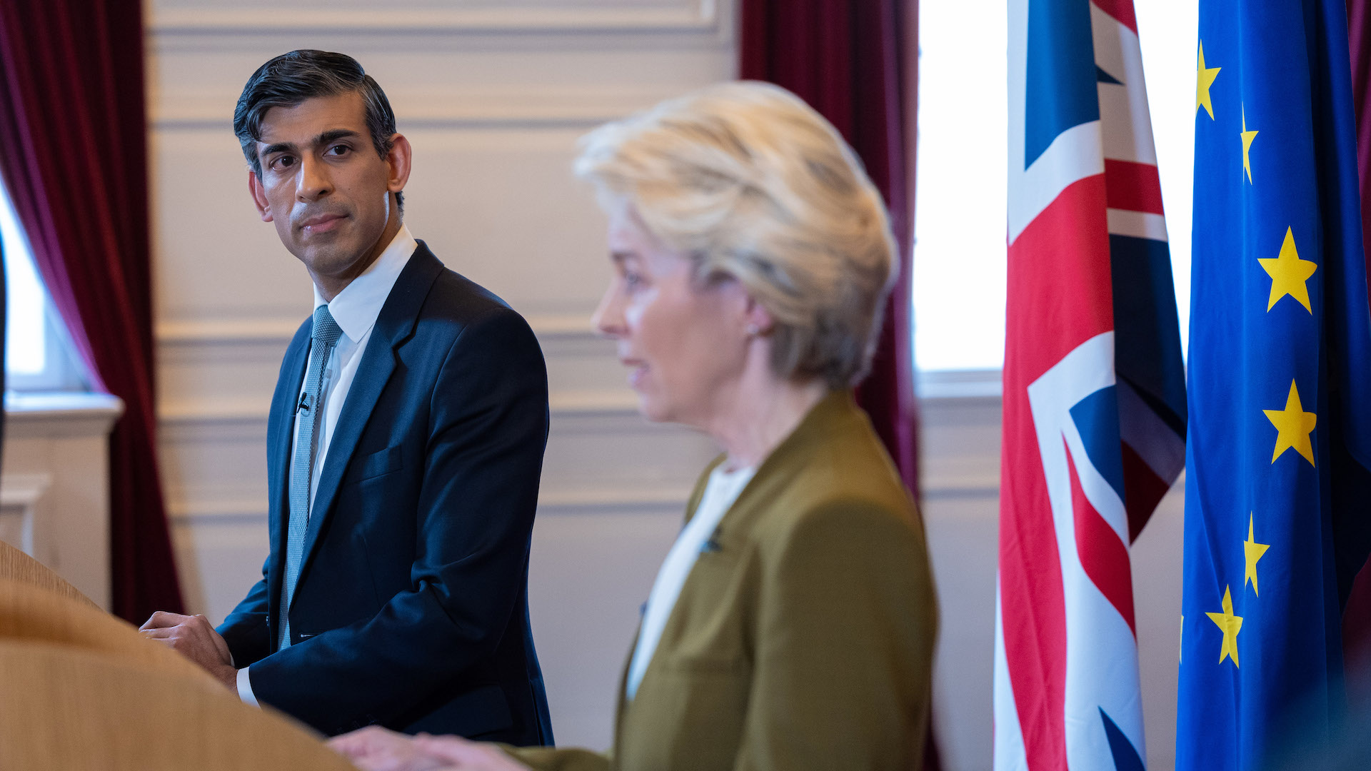 Rishi Sunak stands in front of a lectern and looks at Ursula von der Leyen, who is speaking at her lectern