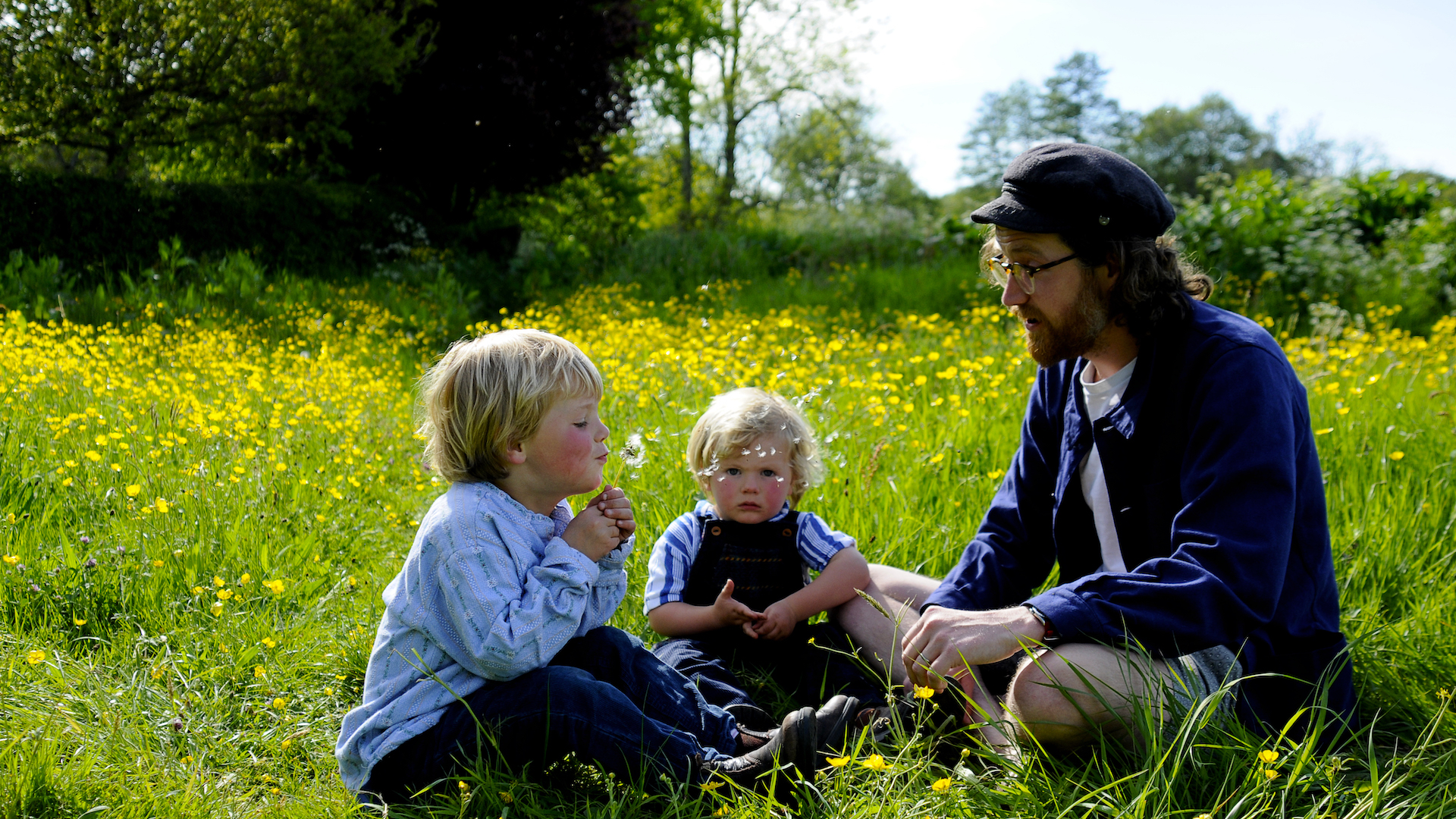 Two red faced children and a man sit in a meadow in summer surrounded by long grass and wildflowers