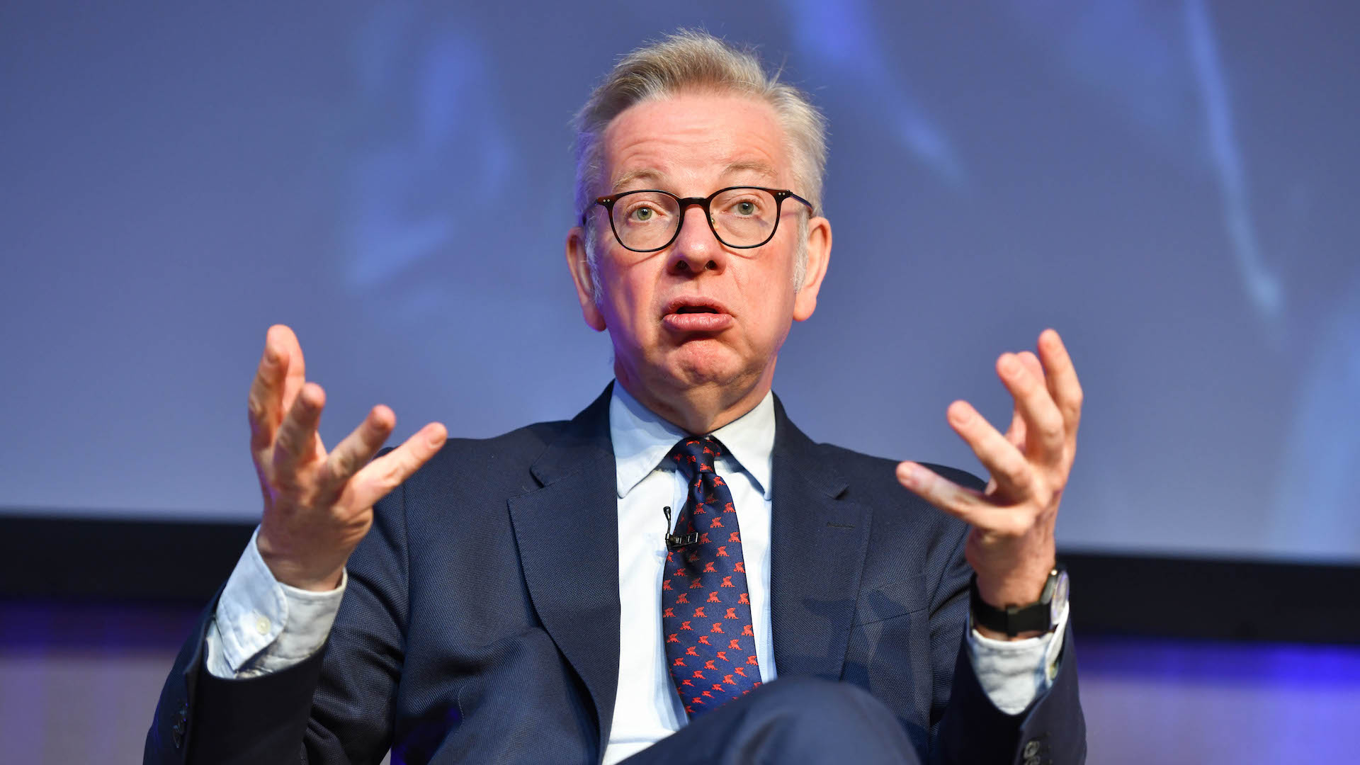 Michael Gove sits in a dark suit, light shirt, dark tie with his hands held out in front of him