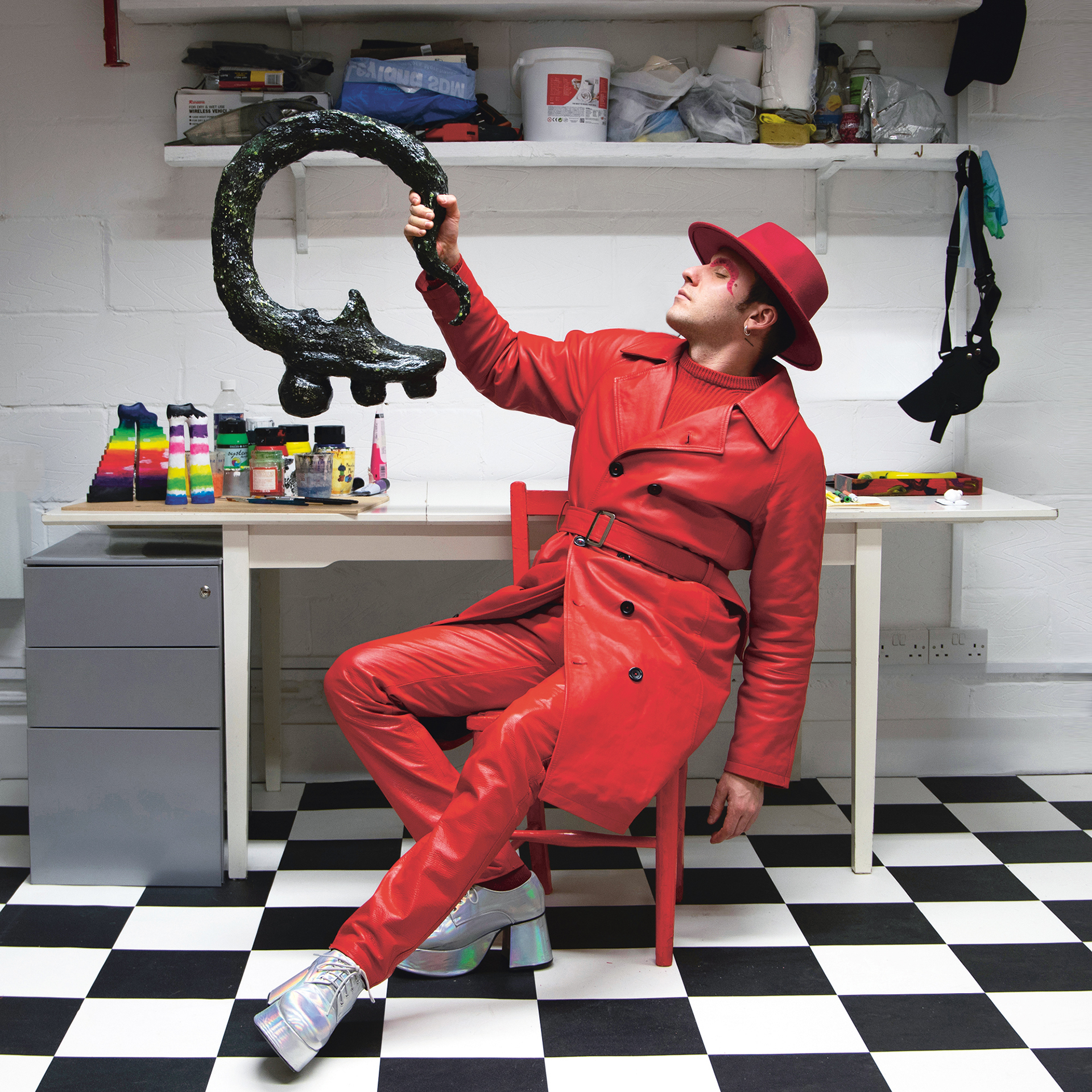 A man dressed in red hat, coat and trousers poses on a chair on a black and white floor. Image: Rose Leahy