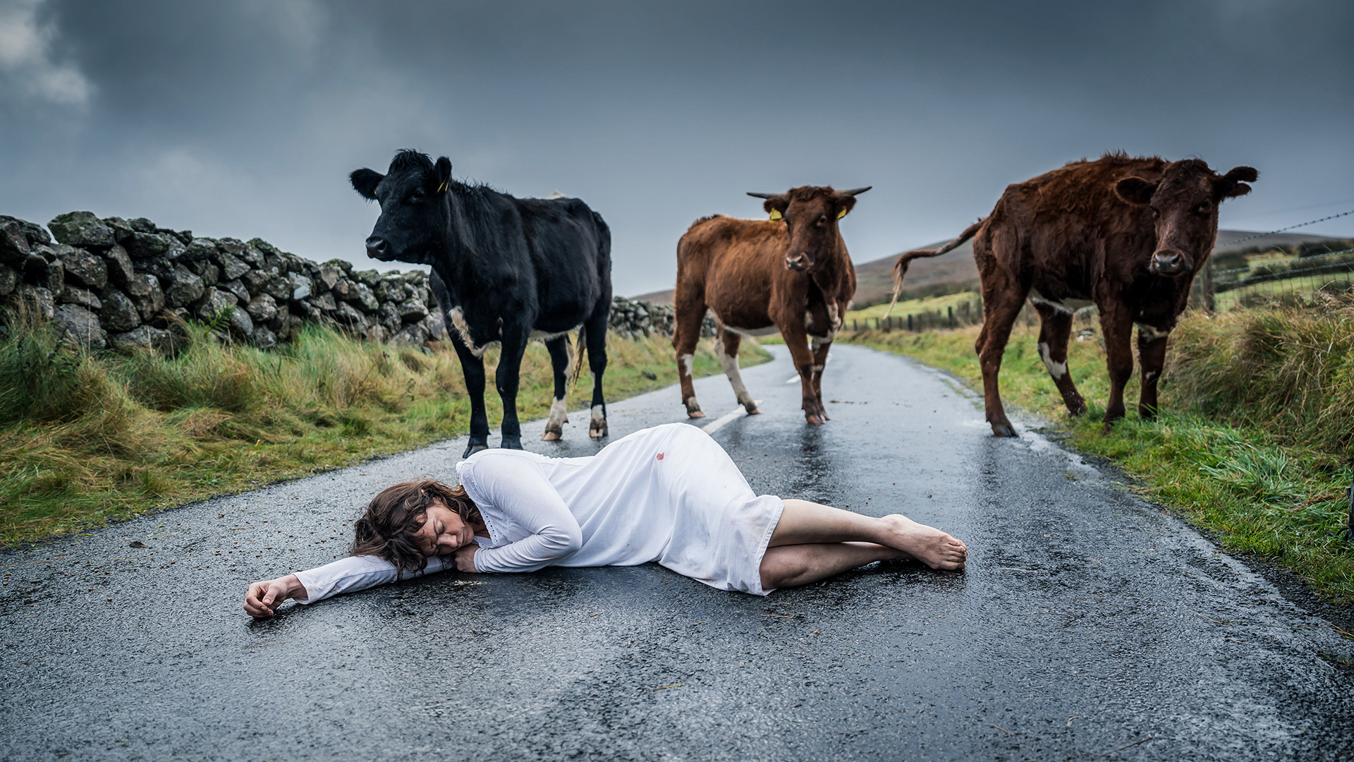 A woman lies on the road in a nightgown surrounded by cows