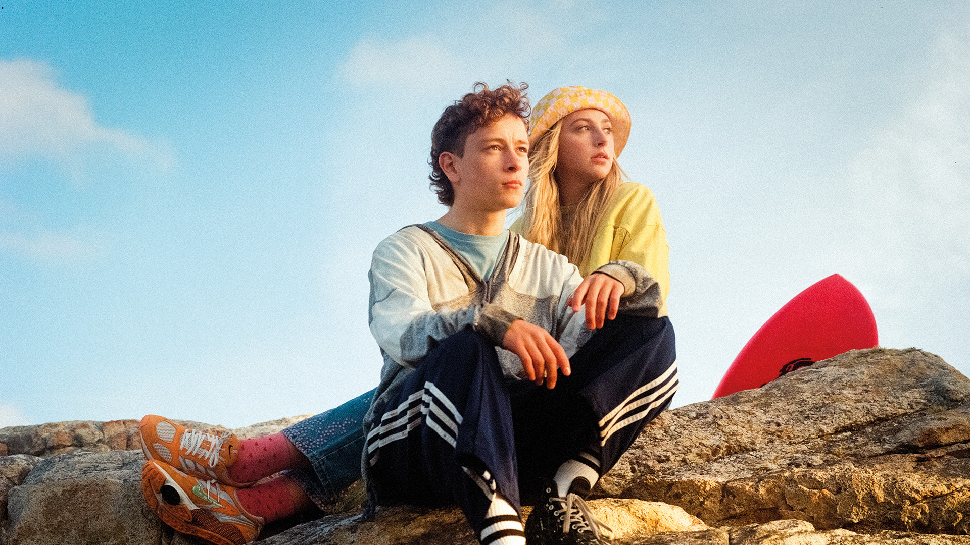 A boy and a girl sit on a rock beside a surfboard