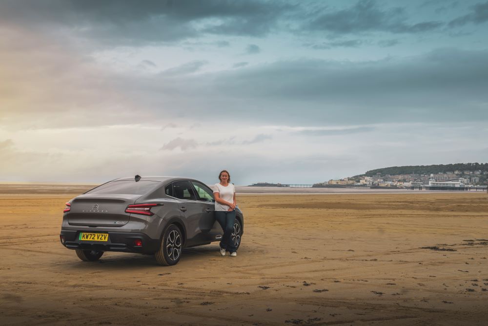 Vicky Parrott leans on Citroën ë-C4 X Electric, which is parked on an empty beach