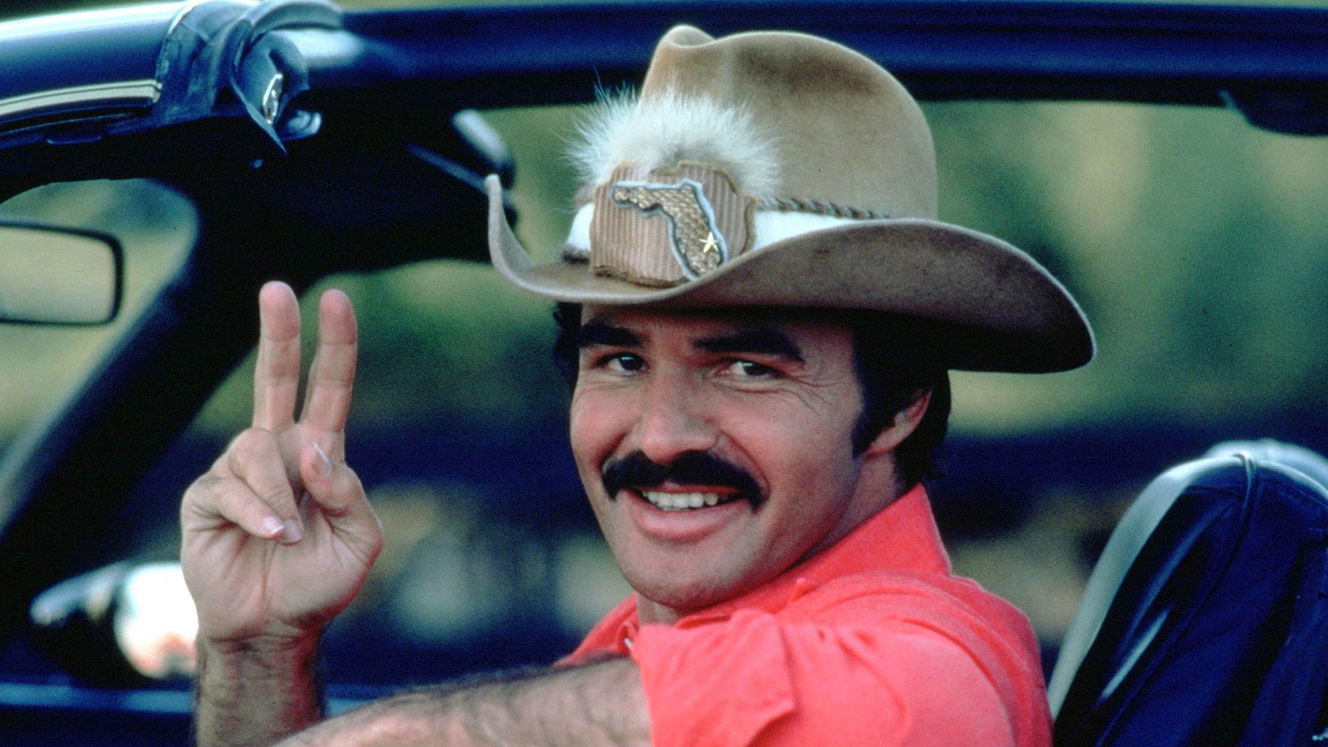Burt Reynolds in a car wearing pink shirt and cowboy hat