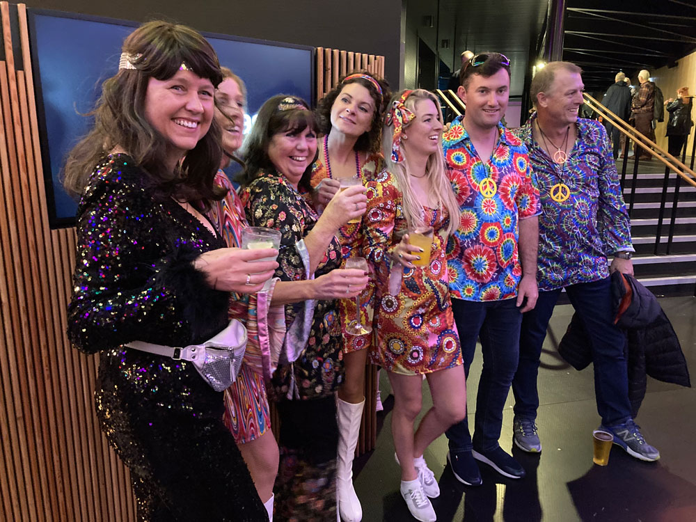 A group of seven men and women in flower power outfits outside ABBA Voyage